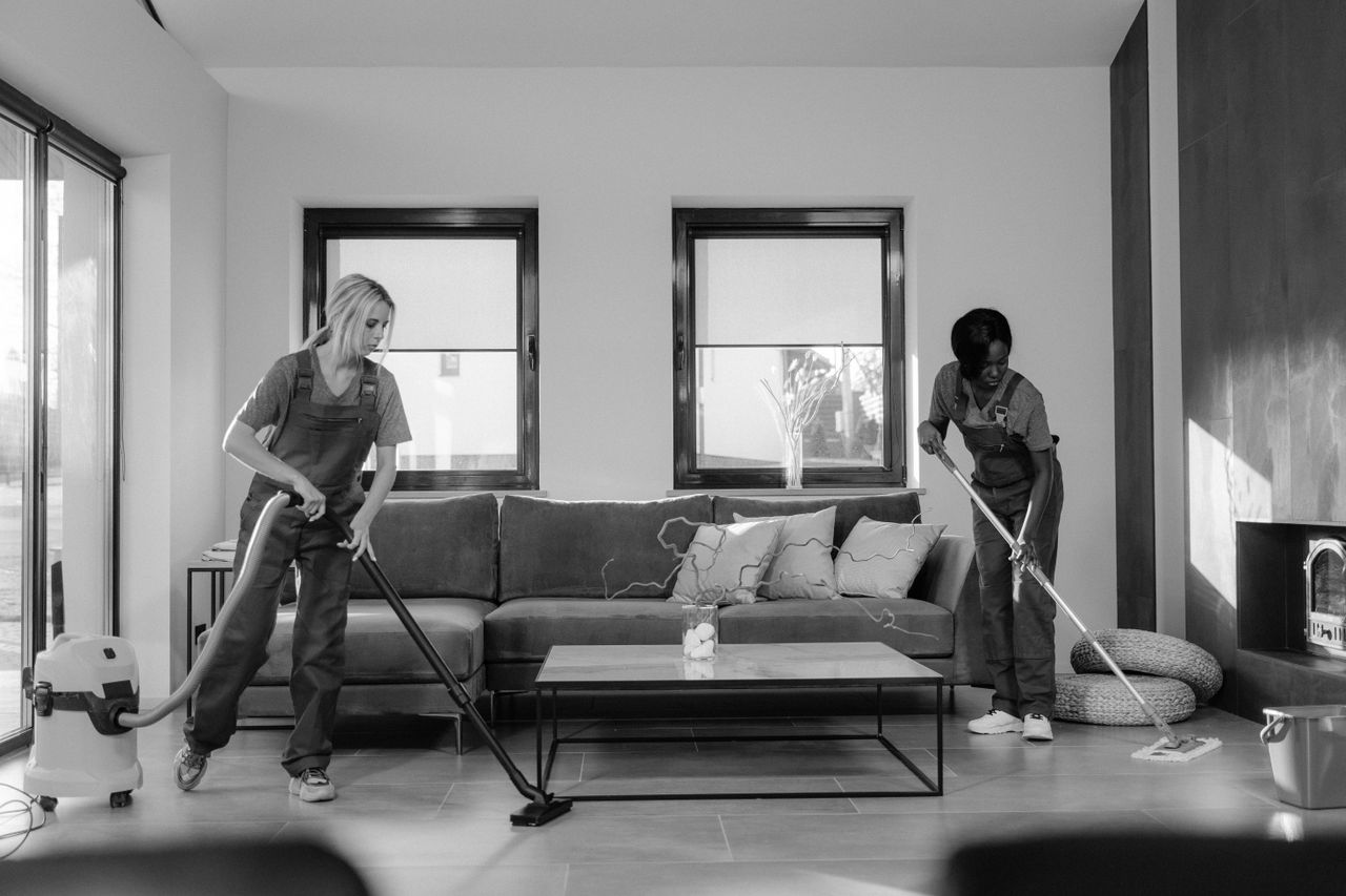 Dve ženy v pracovnom oblečení upratujú modernú obývačku pomocou vysávača a mopu.